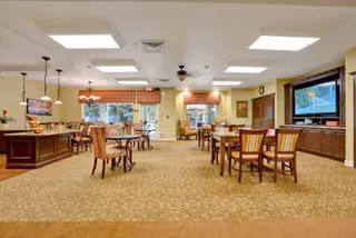 A spacious dining room with multiple wooden tables and chairs arranged neatly. The room has large windows with valances, a carpeted floor, and ceiling lights including pendant lights over a buffet area. A large flat-screen TV is mounted on the wall above a wooden cabinet.