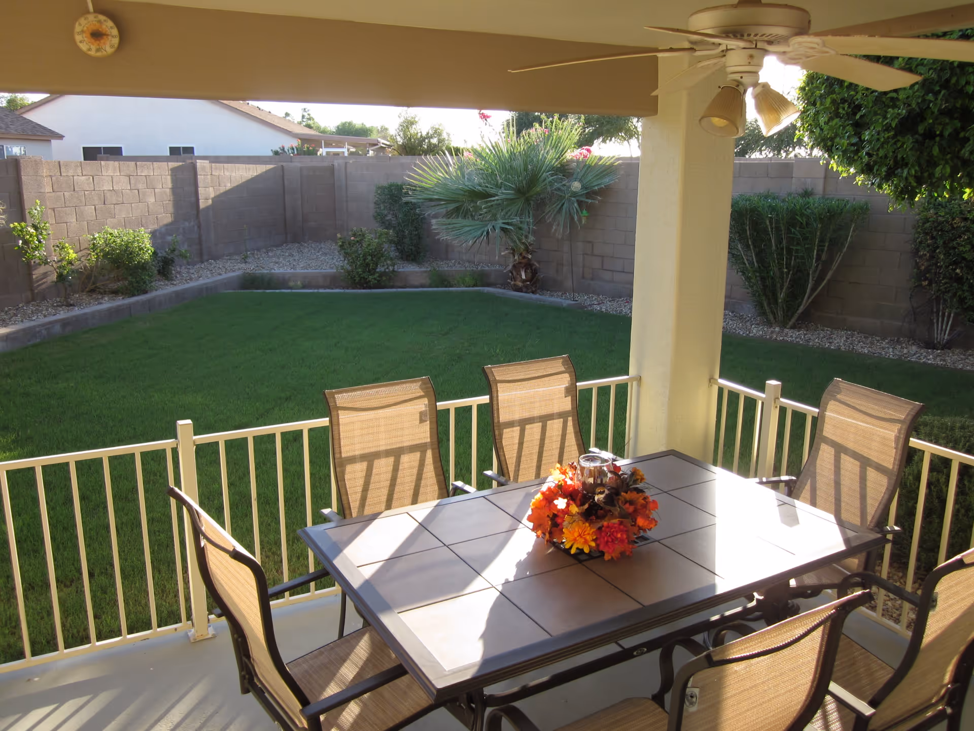 Covered outdoor patio area with a ceiling fan, a rectangular table with a floral centerpiece, and six chairs. The patio overlooks a green lawn with various bushes and a palm tree, enclosed by a brick wall fence.