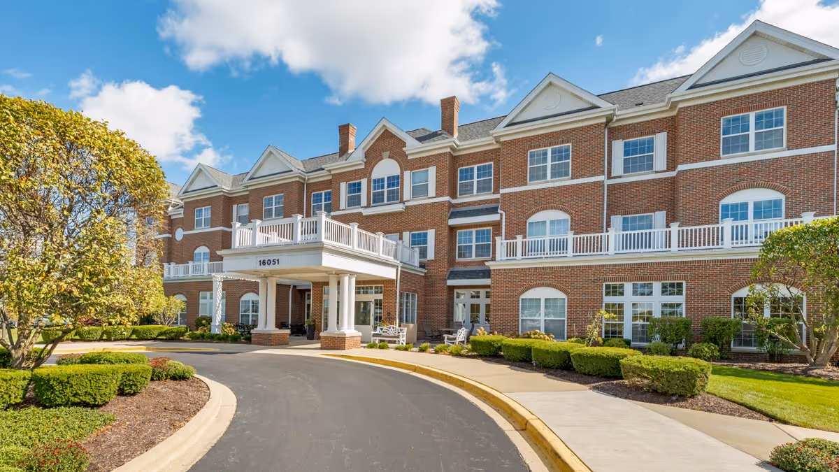 Exterior view of a three-story brick senior living facility with white trim and multiple windows. The entrance features a covered drop-off area with white columns and a balcony above. Well-maintained landscaping with trimmed bushes and trees surrounds the building under a partly cloudy blue sky.