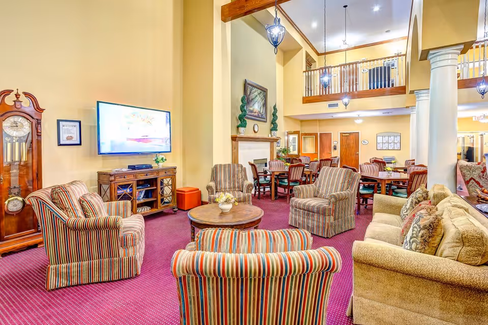 A spacious and well-lit living room area in a senior living facility featuring striped upholstered armchairs arranged around a round wooden coffee table, a beige sofa with decorative pillows, a large flat-screen TV mounted on the wall above a wooden media console, a tall grandfather clock, and a fireplace with a painting and topiary decorations above it. The room has high ceilings with hanging lantern-style lights and a balcony railing overlooking the space. In the background, there are multiple wooden dining tables with chairs.