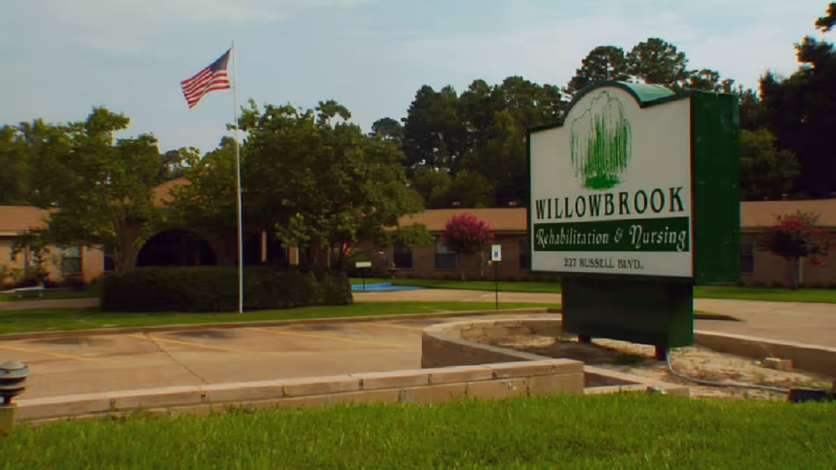 Large green sign reading 'Willowbrook Rehabilitation & Nursing' in front of a single-story nursing center with an American flag, parking area, and landscaping.