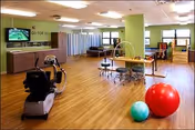 Bright interior rehab gym with exercise equipment, therapy tables, and large windows.