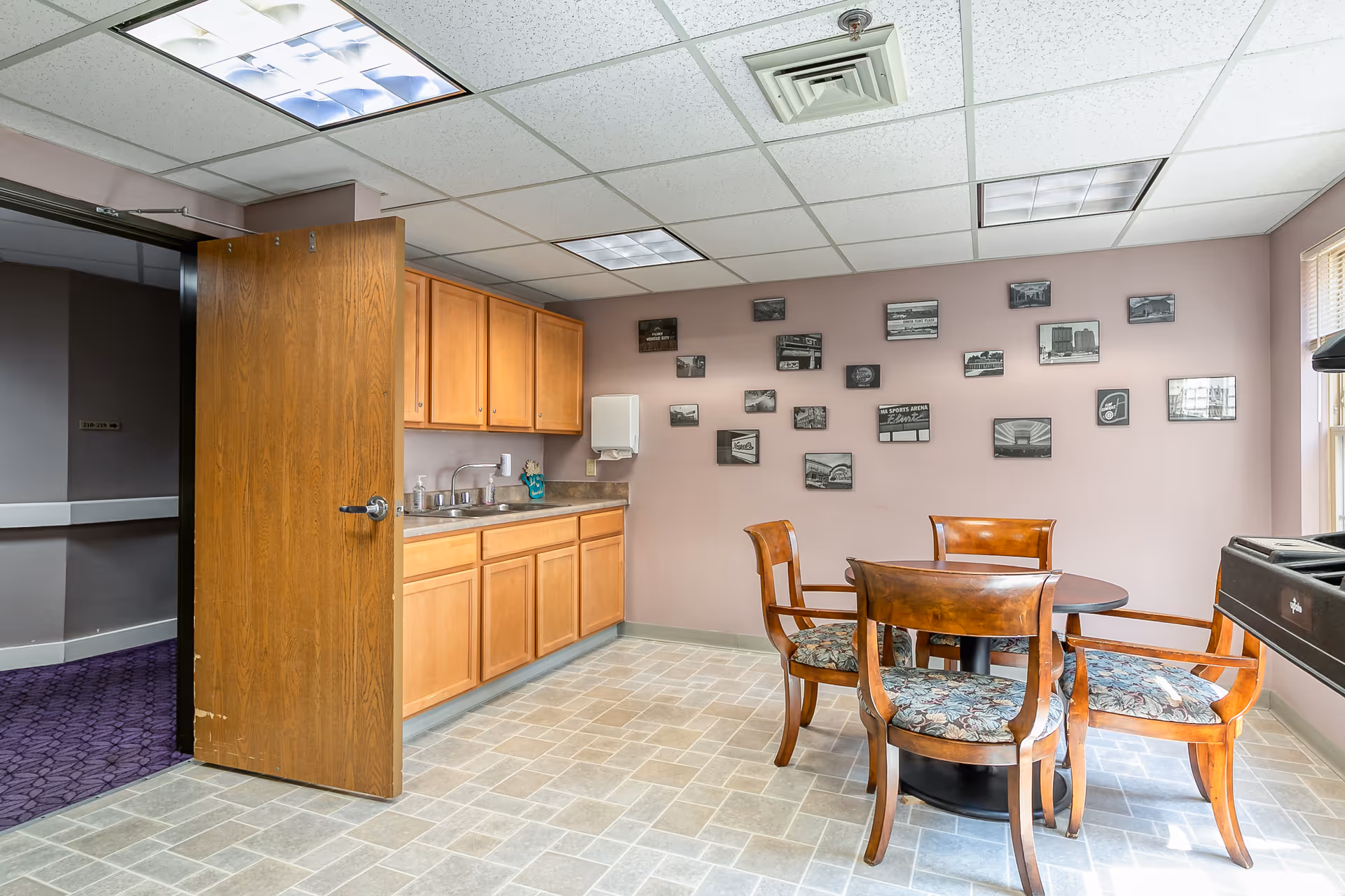 A small dining area in Rosehaven Manor featuring a round wooden table with four wooden chairs that have floral-patterned cushions. The room has light pink walls decorated with multiple framed black and white photos. There is a kitchenette with wooden cabinets, a countertop, a sink, and a paper towel dispenser. The floor is tiled with a light gray pattern, and a wooden door is open to a hallway with purple carpet.