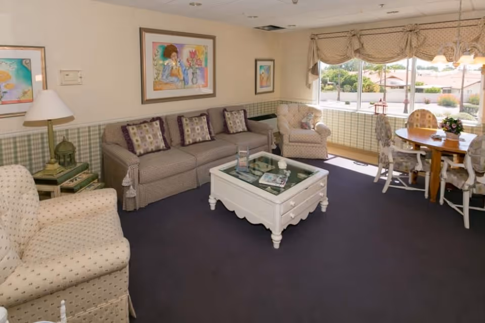 A cozy living room area in a senior living community featuring a beige patterned armchair, a checkered sofa with decorative pillows, a white coffee table with glass top, and a round dining table with four chairs near a large window with a view of the outdoors. The walls have framed artwork and a lamp on a side table.