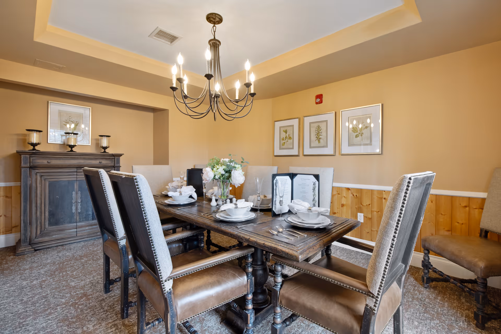 A dining room with a wooden table set for six people, featuring white plates, bowls, napkins, and silverware. The room has beige walls with wooden paneling on the lower half, three framed botanical prints on the wall, a dark wooden sideboard with three candle holders, and a chandelier with lit bulbs hanging from the ceiling.