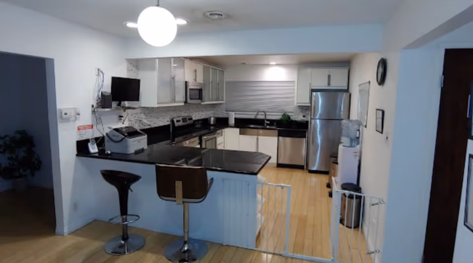 Modern kitchen with black countertops, white cabinets, stainless steel appliances including a refrigerator, microwave, and dishwasher. Two bar stools are placed at the kitchen island. A water cooler and a clock are visible on the right wall. The floor is light wood, and a baby gate is installed at the entrance to the kitchen area.
