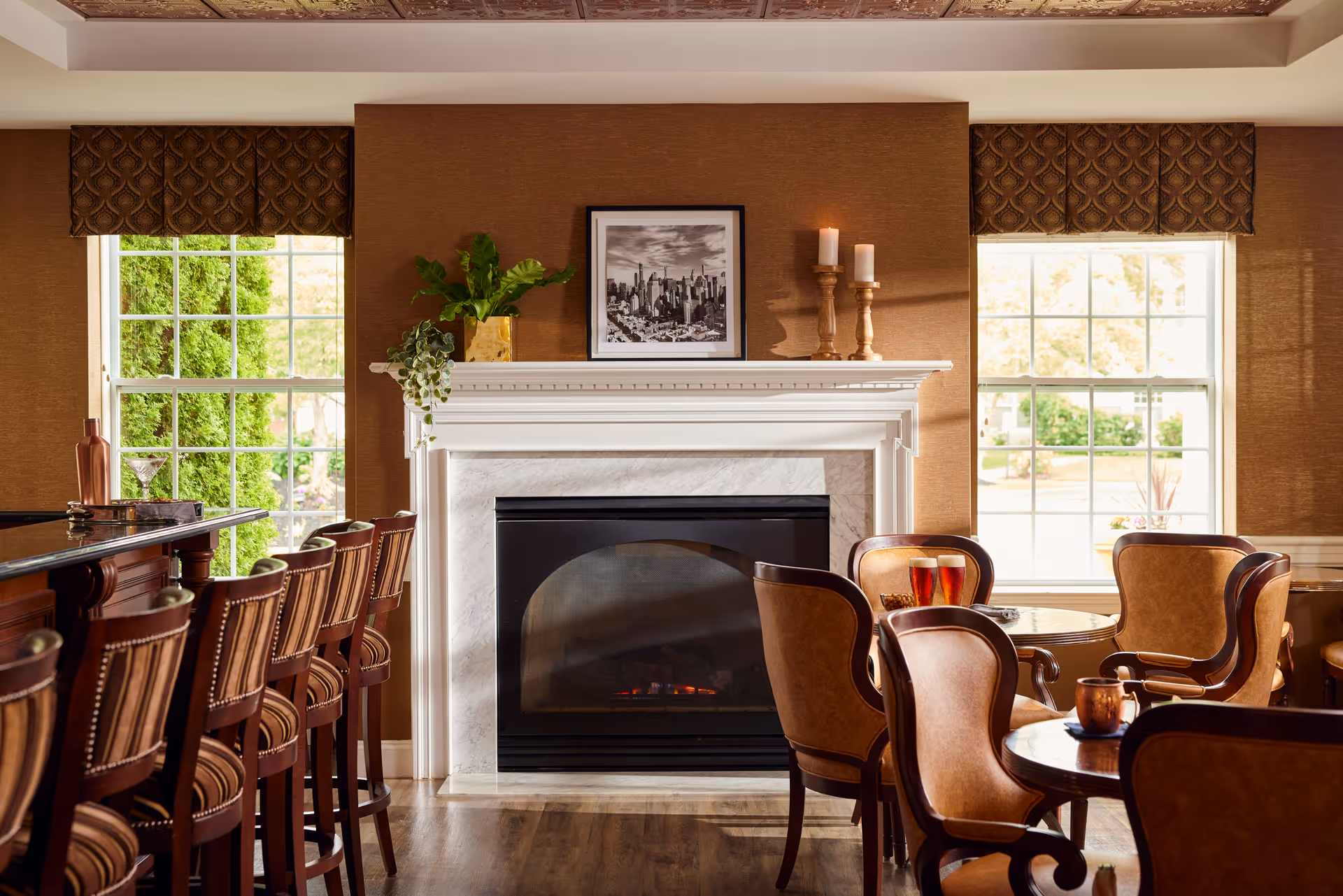 A cozy lounge area with a white fireplace in the center, decorated with a black and white cityscape photo, two candles, and green plants. To the left, there is a bar counter with several wooden bar stools. To the right, there are round tables surrounded by cushioned chairs. Two large windows with brown valances let in natural light and show greenery outside.