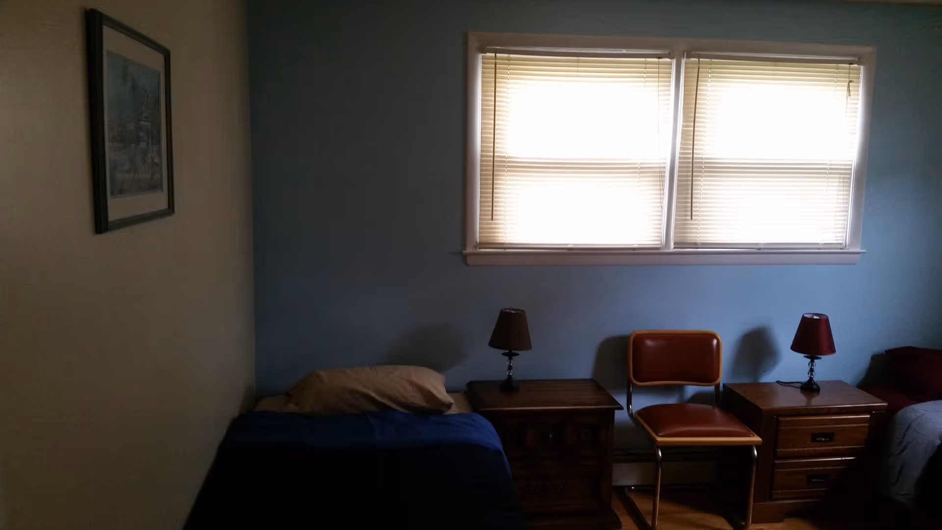 Simple bedroom with two single beds, wooden nightstands and lamps beneath a pair of blinds-covered windows.