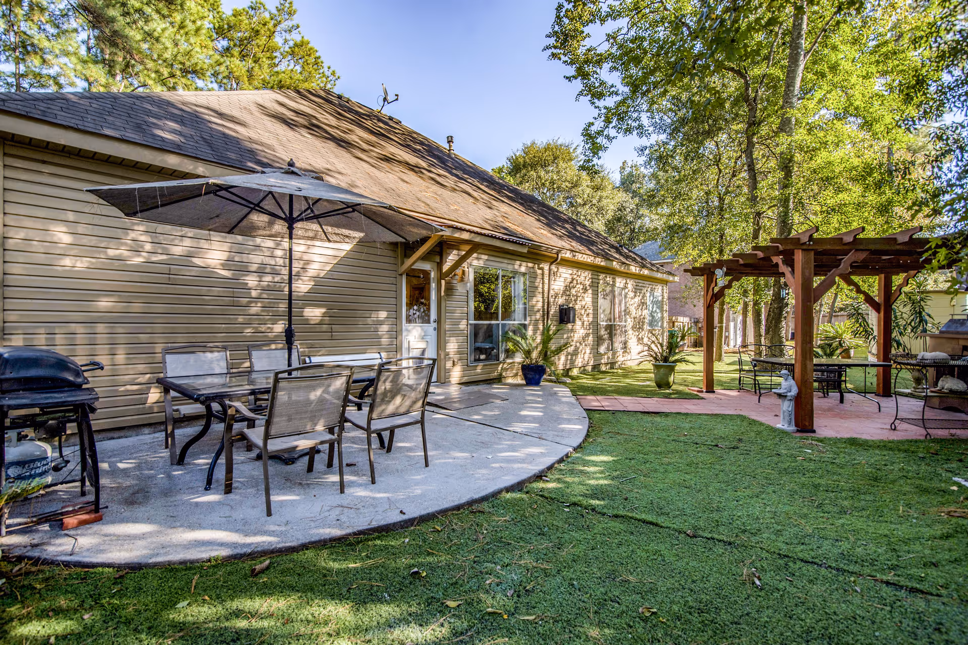 Outdoor patio area at Acorn Manor Assisted Living featuring a concrete seating area with a table, six chairs, and a large umbrella. There is a barbecue grill on the left side. To the right, there is a wooden pergola with additional seating underneath. The area is surrounded by green grass and trees, with the building's beige siding and windows visible in the background under a clear blue sky.
