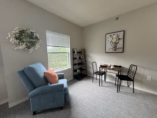 A small living area with a blue upholstered armchair with a red pillow, a window with blinds, a black shelving unit with decorative items, and a small wooden table with two black chairs. A floral painting hangs on the wall above the table, and a decorative wreath with the word 'Welcome' is on the wall near the window.