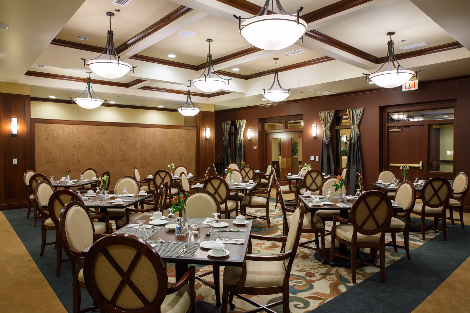 A well-lit dining room with multiple tables set with white cups, saucers, glasses, and silverware. Each table has a small flower arrangement. The room features wooden paneling, patterned carpet, and ceiling lights with decorative fixtures. There are windows with curtains and double doors at the back.