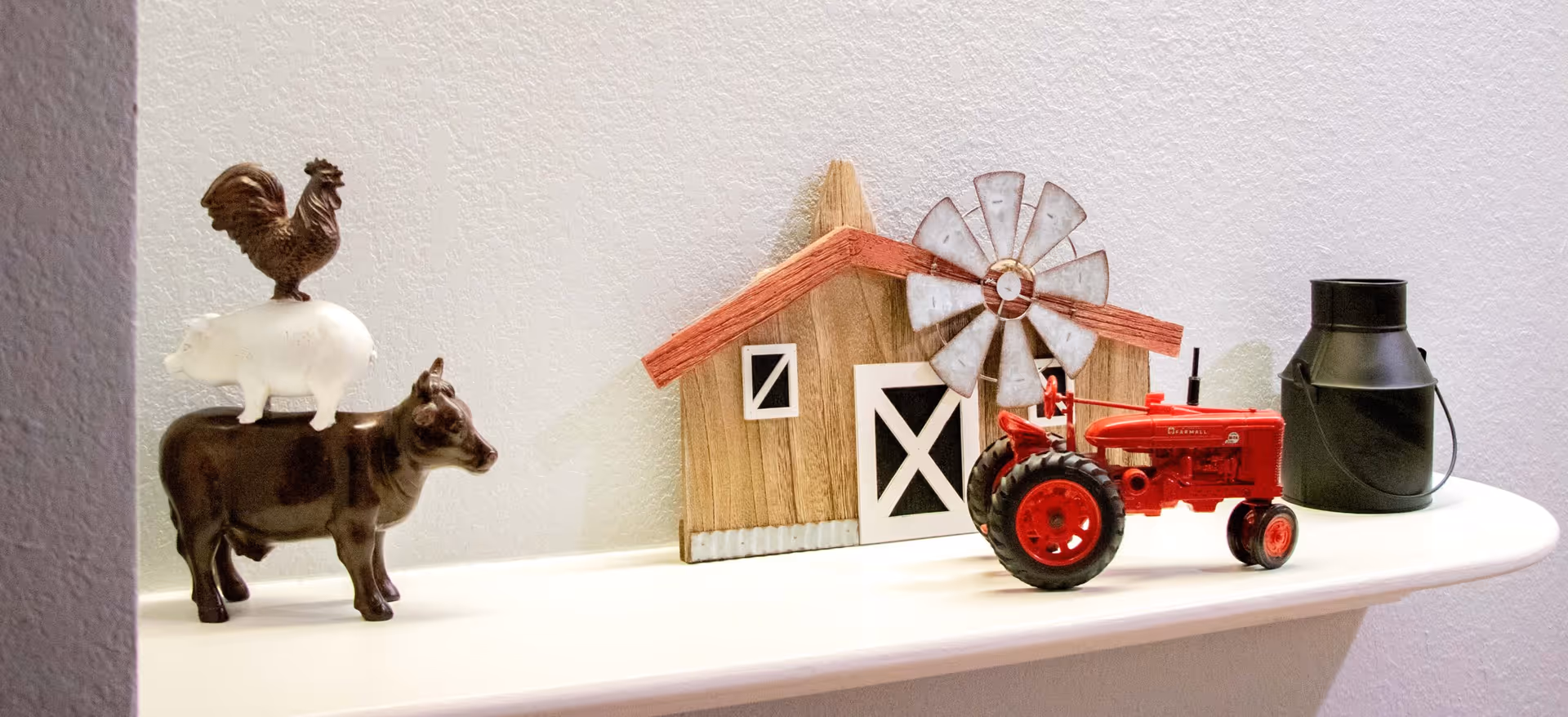A decorative shelf displaying a small stack of ceramic farm animals including a cow, pig, and rooster, a miniature wooden barn with a windmill, a red toy tractor, and a small black metal milk can.