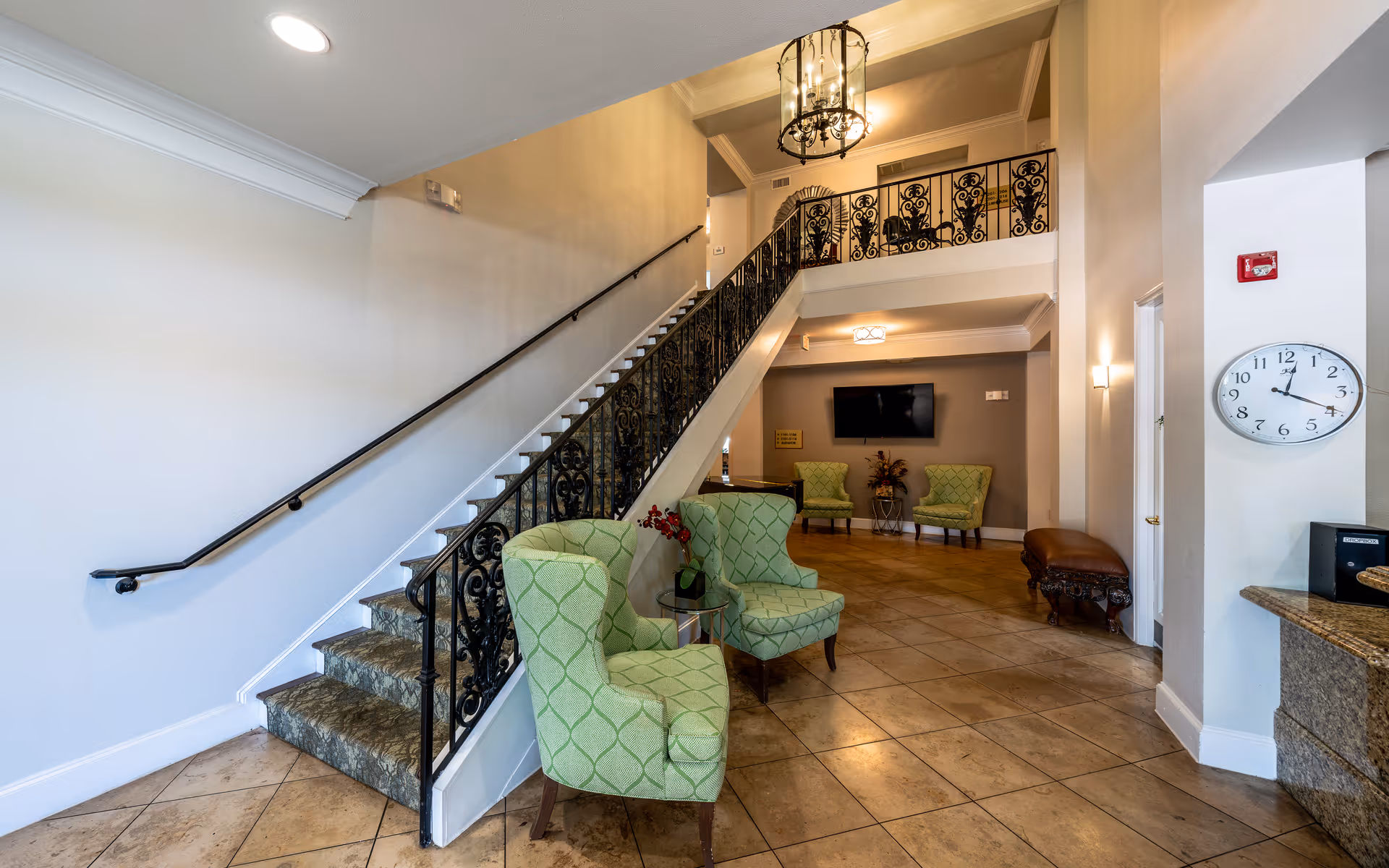 Interior view of a senior living facility lobby with a staircase featuring ornate black railings. Two green patterned armchairs are placed near the base of the stairs, and additional seating with two more green chairs is visible in the background. The floor is tiled, and a wall clock shows the time as 3:15. A flat-screen TV is mounted on the far wall, and a chandelier hangs from the ceiling above the staircase.