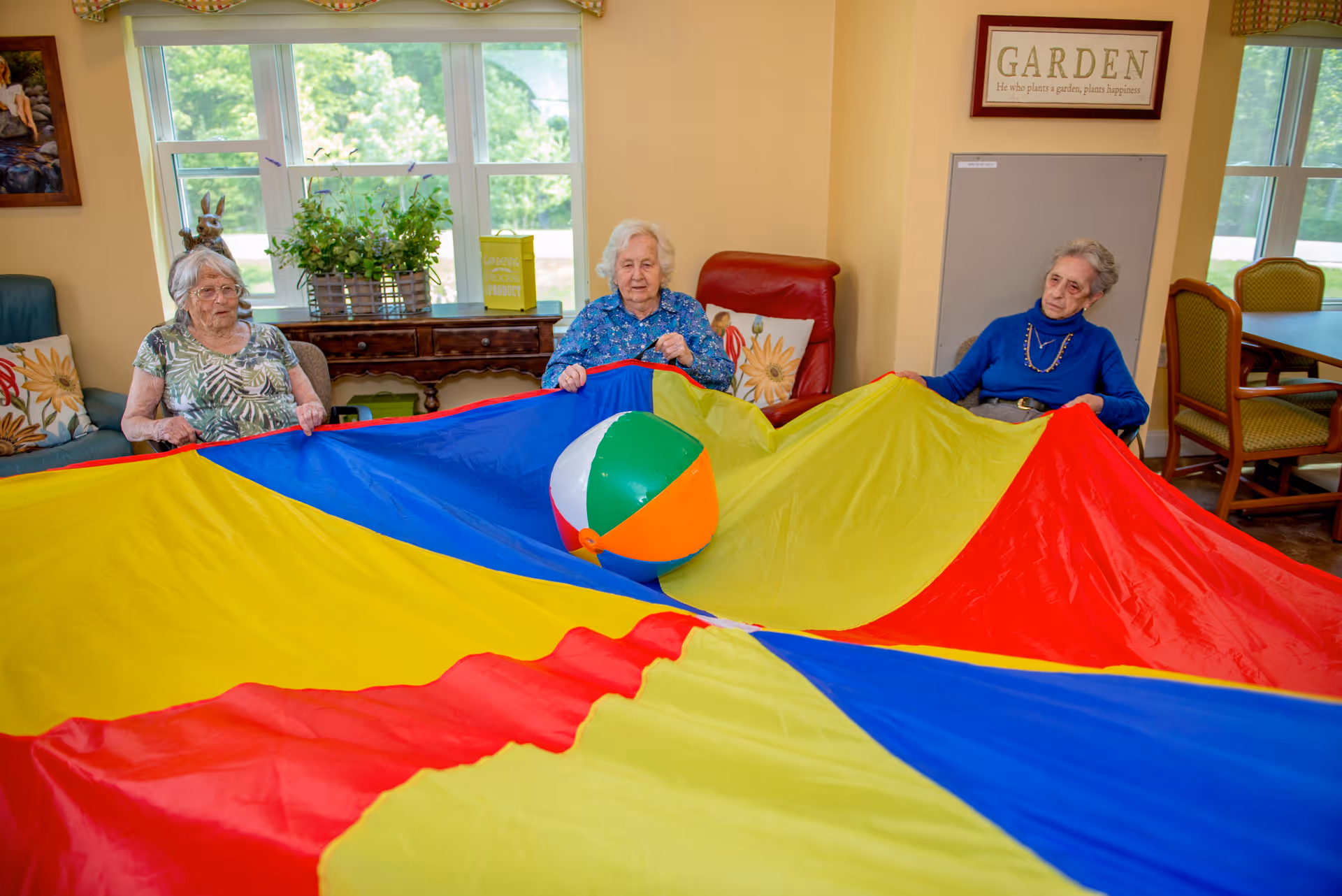 Three elderly women sitting in a bright room holding the edges of a large colorful parachute with a beach ball on top of it. The room has large windows, plants on a wooden table, and a framed sign on the wall that says 'GARDEN'.