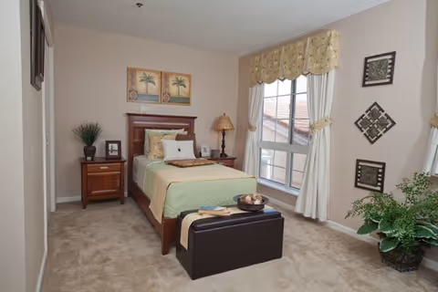 A neatly arranged bedroom with a single bed featuring green and beige bedding, a wooden headboard, and a nightstand with a lamp and decorative items. There is a window with cream-colored curtains and valance, wall art above the bed and on the adjacent wall, a small bench at the foot of the bed, and a potted plant in the corner.