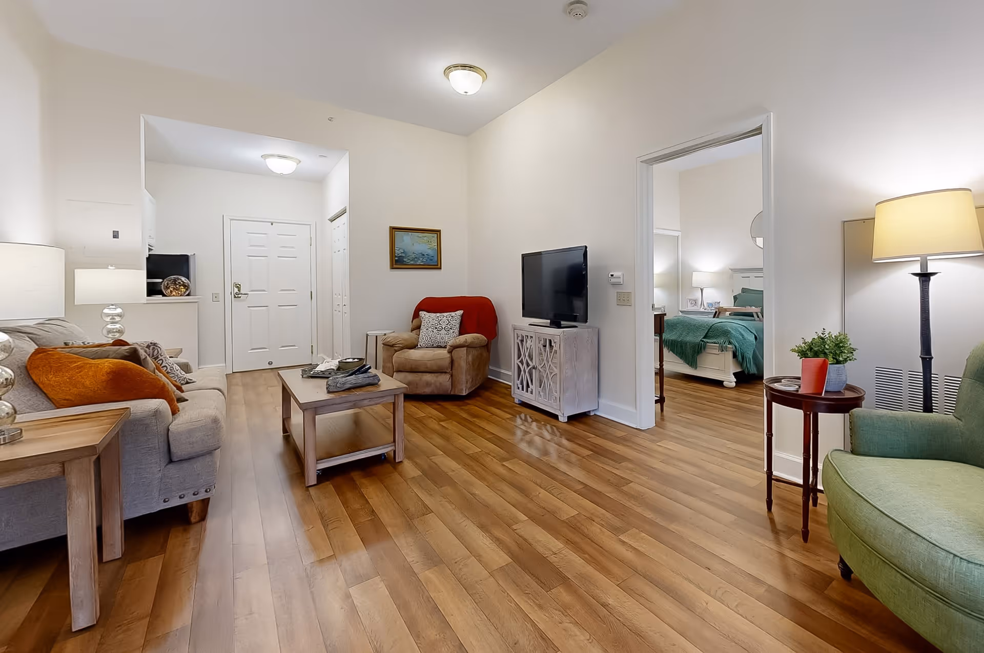 A cozy living room with wooden flooring, a beige sofa with orange and patterned cushions, a beige armchair with a red throw, a wooden coffee table, and a TV on a white cabinet. A green armchair and a small round table with a lamp and plant are on the right. The room opens to a bedroom with a bed covered in green bedding. The entrance door and a small kitchen area are visible in the background.