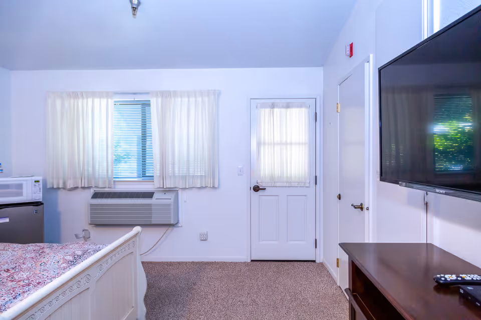 Interior view of a senior living bedroom featuring a bed with a patterned bedspread, a window with sheer curtains and blinds, an air conditioning unit below the window, a white door with a sheer curtain, a wall-mounted flat-screen TV, and a wooden dresser with a remote control on top.