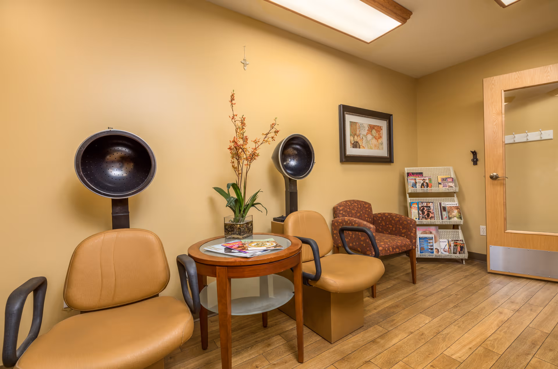 Interior of a hair salon area with two vintage hair dryers attached to tan chairs, a small round wooden table with magazines and a plant, a patterned armchair, a magazine rack, and a wooden door with a glass panel. The walls are painted beige and the floor is wooden.