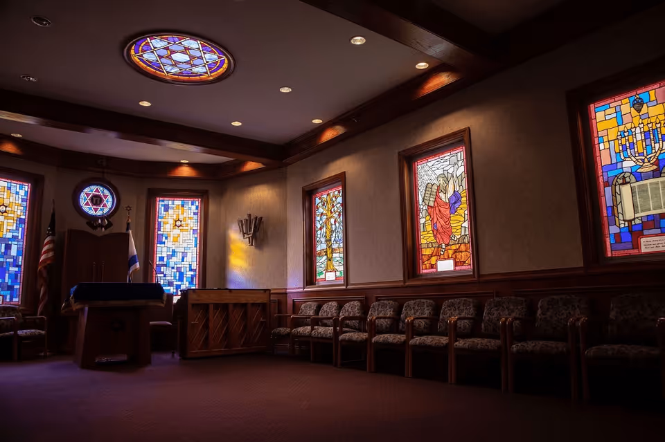 Interior of a room with stained glass windows depicting religious symbols and scenes, a row of cushioned chairs along the wall, a wooden podium with a blue cloth, an upright piano, and flags in the corner. The ceiling has recessed lighting and a circular stained glass light fixture.