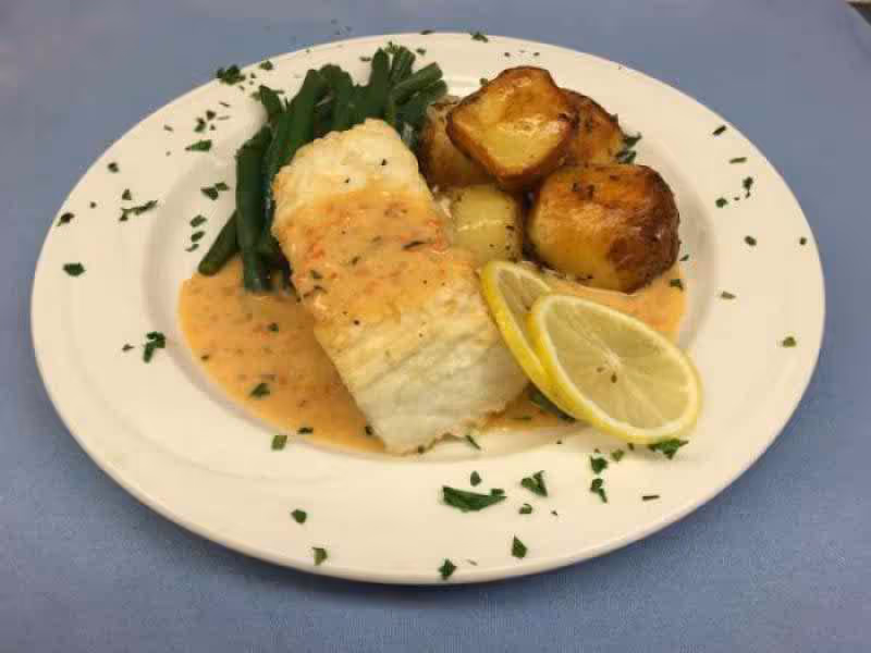 A plate with a piece of white fish topped with sauce, green beans, roasted potatoes, and lemon slices on a blue tablecloth.