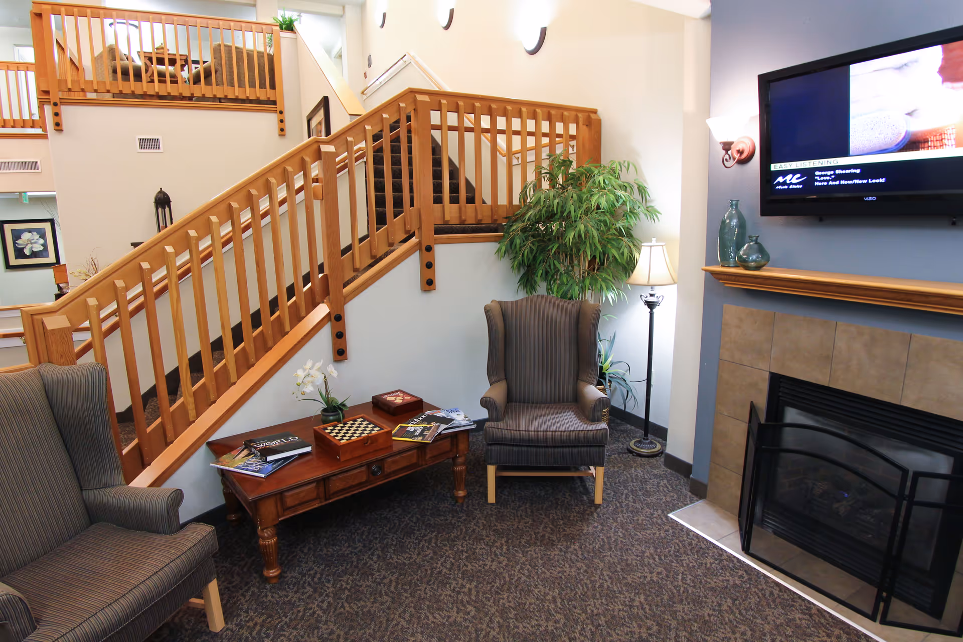 A cozy living room area with two striped armchairs, a wooden coffee table with books and a chessboard, a potted plant, a floor lamp, a fireplace with a tiled surround, and a wall-mounted TV above the fireplace. A wooden staircase with railings is visible in the background.