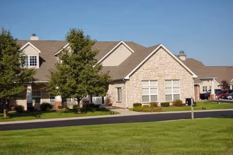 Exterior view of a single-story senior living facility building with beige stone and siding, multiple windows, a well-maintained lawn, and a paved driveway under a clear blue sky.