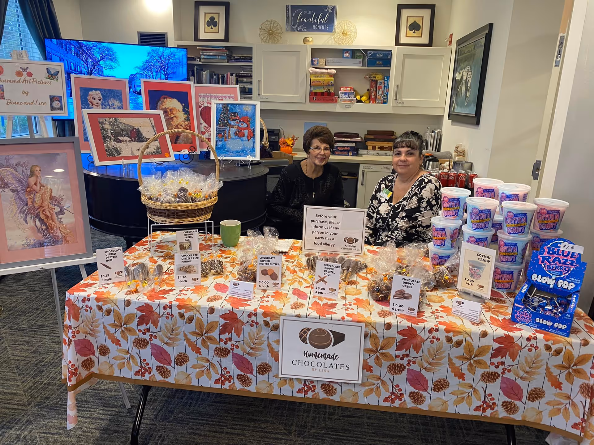 Two women sitting behind a table covered with a fall-themed tablecloth displaying homemade chocolates, cotton candy, and blue razz berry blow pops for sale. Behind them are shelves with board games and framed artwork displayed on easels and a piano.