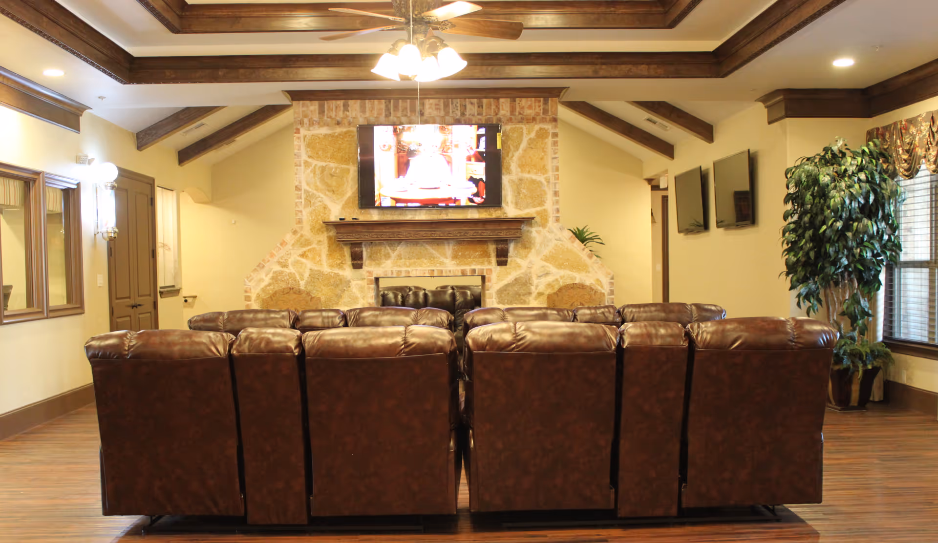 A cozy living room with a row of brown leather recliners facing a stone fireplace with a mounted flat-screen TV above it. The room has wooden ceiling beams, a ceiling fan with lights, wooden floors, and large windows with curtains on the right side. There are also two additional wall-mounted TVs and a large potted plant near the windows.