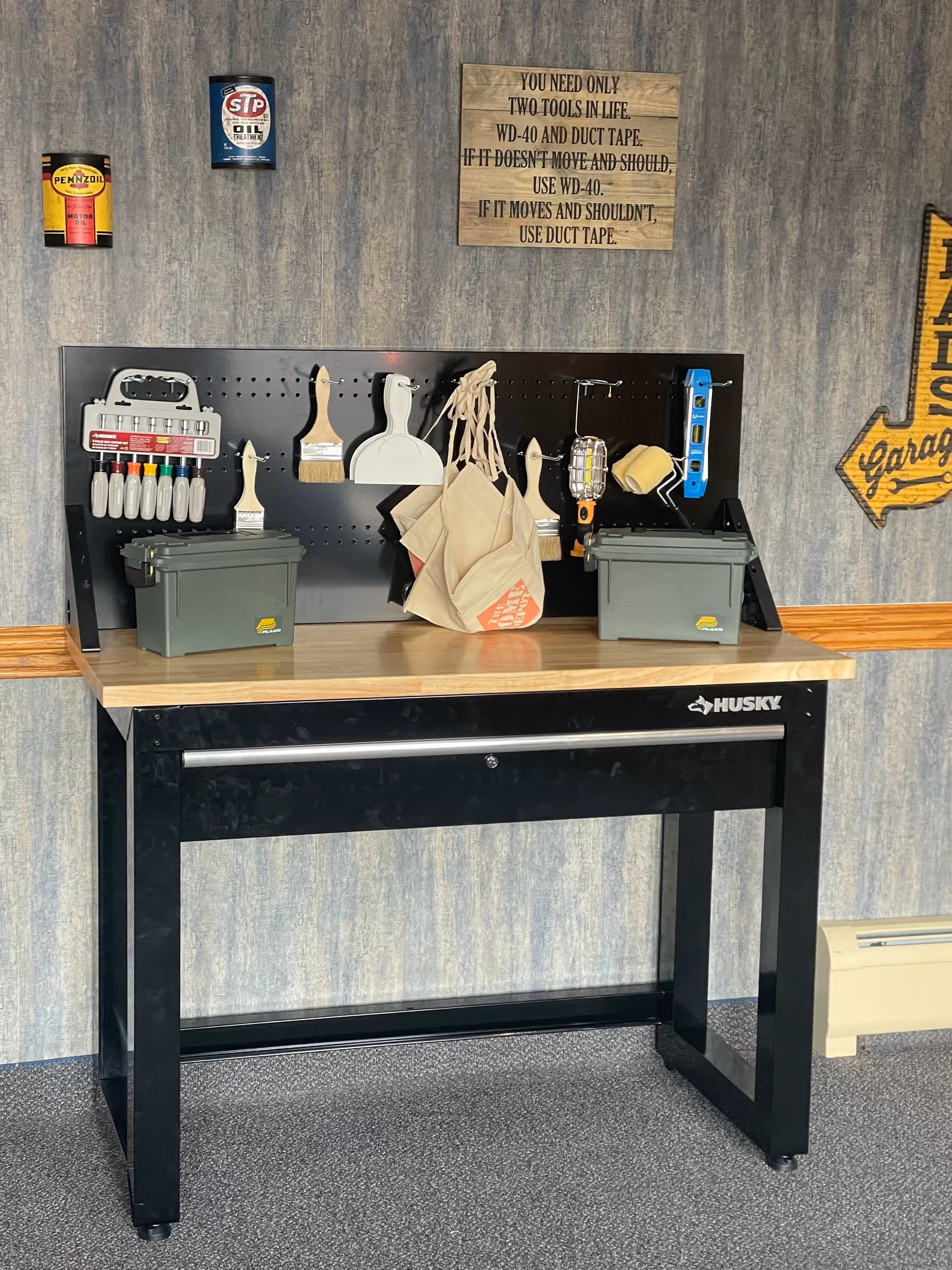 A black Husky workbench with a wooden top and pegboard organizer holding paint brushes, tools, bags and storage boxes beneath a humorous WD-40 and duct tape sign on a wallpapered wall.