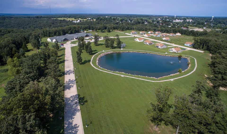 Aerial view of Victorian Place of Owensville showing a large pond with a fountain surrounded by green lawns, a paved road lined with trees, and multiple small residential buildings with orange roofs. The area is surrounded by dense trees and open land under a partly cloudy sky.