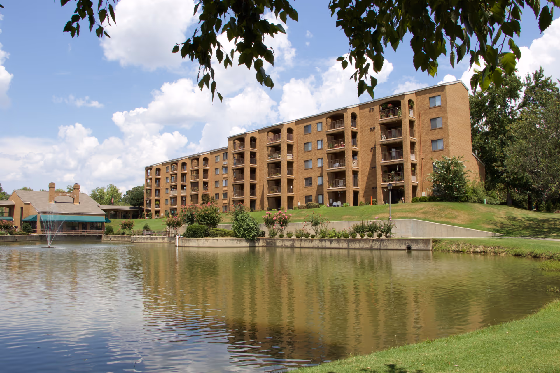 A multi-story brick senior living building with balconies overlooking a pond and fountain under a partly cloudy sky.