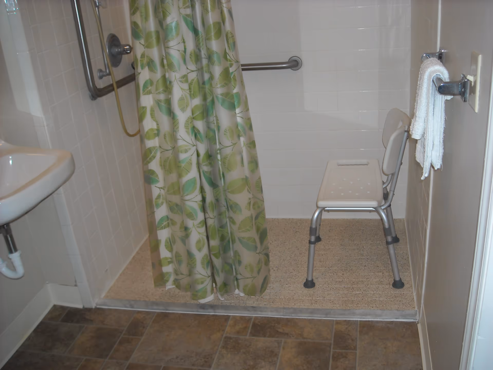 A bathroom with a walk-in shower featuring a green and white leaf-patterned shower curtain, a white shower chair with back support, a grab bar on the tiled wall, a white towel hanging on a wall-mounted towel rack, and a small white sink on the left side.