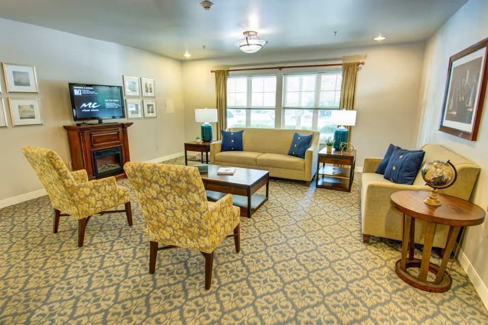 A cozy living room with two beige sofas and two yellow patterned armchairs arranged around a wooden coffee table. The room has a patterned carpet, a large window with curtains, two side tables with lamps, a small round table with a globe, framed pictures on the walls, and a TV on a wooden stand.