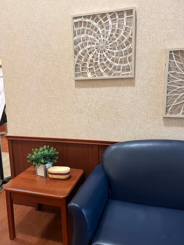 A corner of a room with a dark blue leather armchair next to a wooden side table. On the table, there is a small green potted plant and a small decorative box. The wall behind has a beige patterned wallpaper and two framed decorative wall art pieces with a spiral design.