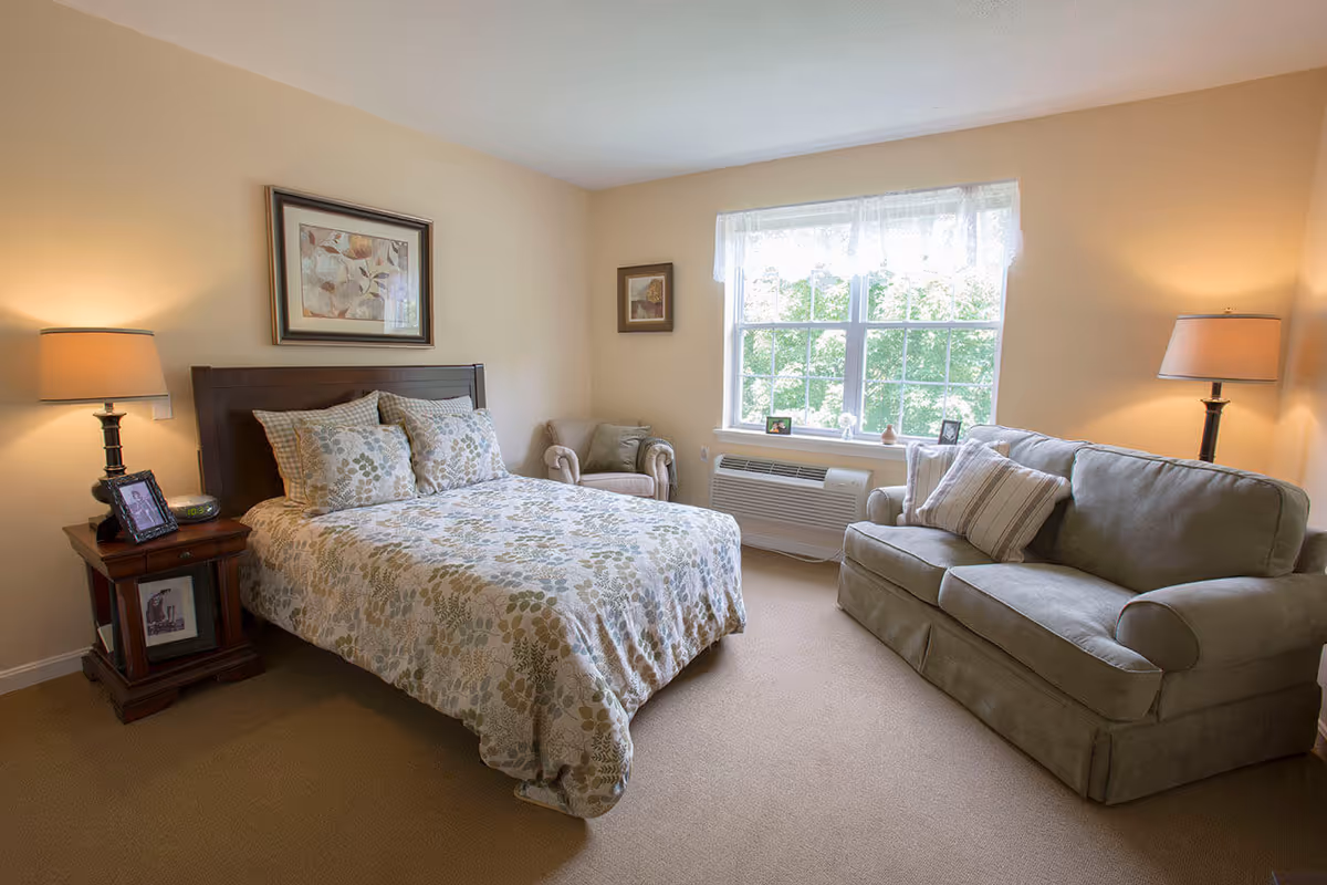 Sunlit furnished bedroom with a bed, nightstand, armchair, and a sofa by a large window.