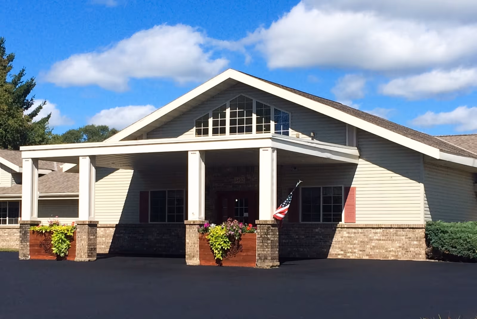Front exterior view of a single-story senior living facility building with a covered entrance supported by white columns, brick and siding exterior, flower planters, an American flag, and a bright blue sky with some clouds.