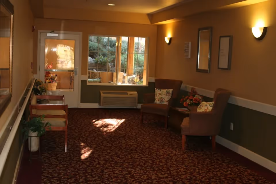 A cozy hallway area in an assisted living facility with patterned carpet, two upholstered armchairs with floral cushions, a small table with a flower arrangement, wall-mounted lights, a window showing an outdoor patio, and a door at the end of the hallway.