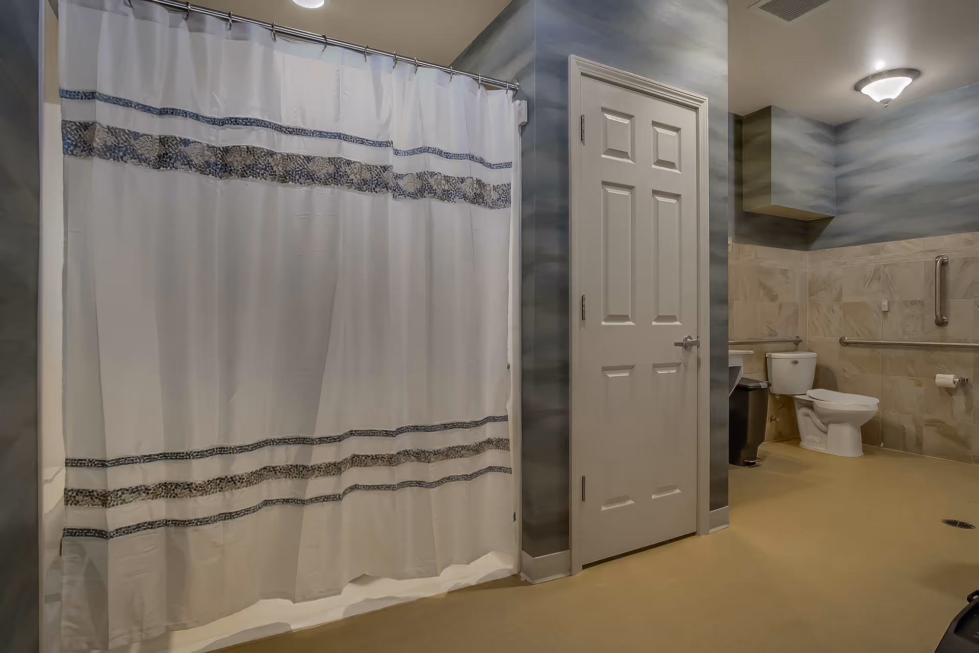 A spacious bathroom with a white shower curtain featuring blue and beige decorative stripes, a closed white door, a toilet with grab bars on the tiled walls, a trash can, and a ceiling light fixture.