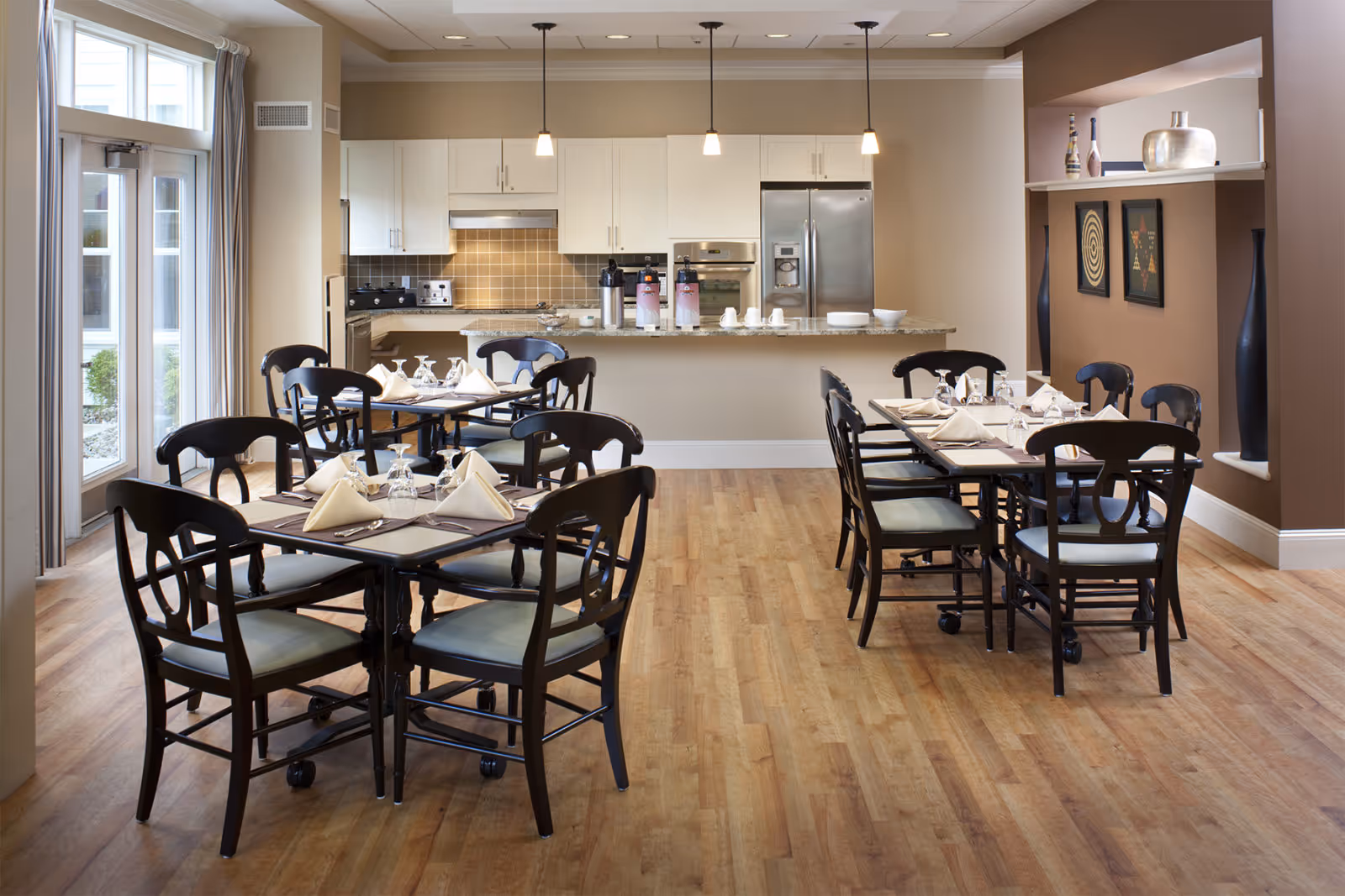 A dining area with two tables set for meals, each surrounded by black wooden chairs with light-colored cushions. The tables are neatly arranged with folded napkins, glasses, and silverware. In the background, there is a kitchen area with white cabinets, a stainless steel refrigerator, oven, and a countertop with coffee dispensers. The room has wooden flooring, beige walls, and large windows letting in natural light.
