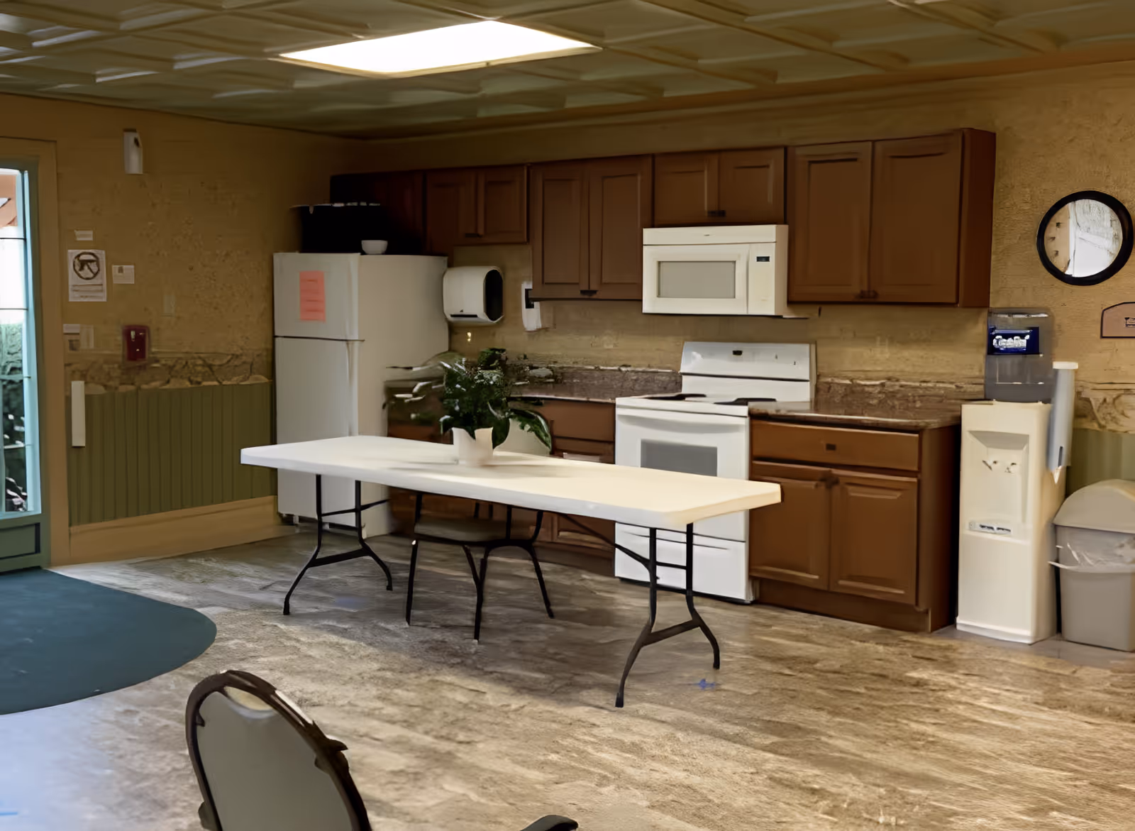 Interior view of a kitchen area with brown cabinets, a white refrigerator, white microwave, and white stove. A long white folding table with a potted plant is placed in the center of the room. There is a water cooler and trash can on the right side, and a door with glass panels on the left side.