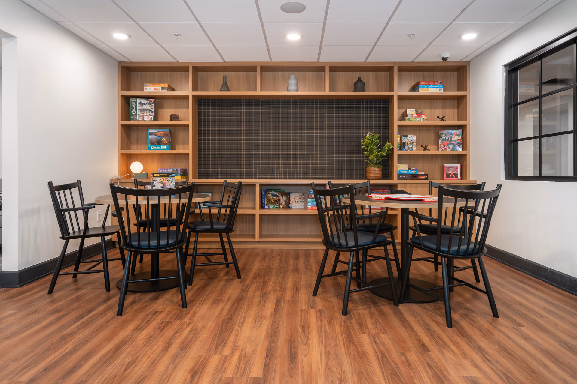 A well-lit room with wooden flooring featuring two round tables surrounded by black chairs. Behind the tables is a large wooden shelving unit filled with board games, puzzles, decorative items, and a potted plant. The room has a modern ceiling with recessed lighting and a window on the right wall.