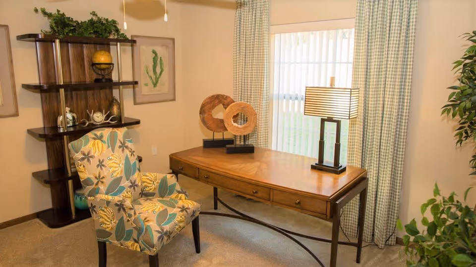 A cozy interior room featuring a wooden desk with three drawers, a decorative table lamp, and two circular wooden sculptures on top. Next to the desk is a floral-patterned upholstered armchair. Behind the desk is a window with vertical blinds and checkered curtains. To the left, there is a dark wooden shelving unit with various decorative items and framed botanical artwork on the wall. A green potted plant is partially visible on the right side of the image.