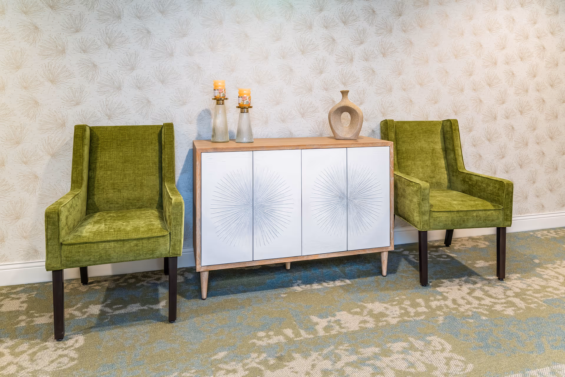 Two green upholstered chairs flank a decorative white cabinet topped with vases against patterned wallpaper.