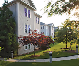 Exterior view of a senior living facility building with multiple windows, surrounded by green grass, trees, and a paved walkway under a clear sky.