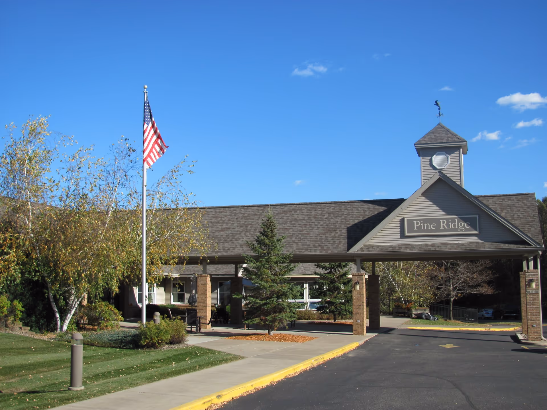 Exterior front view of Pine Ridge Assisted Living facility with a covered entrance, an American flag on a flagpole, trees, and a clear blue sky.