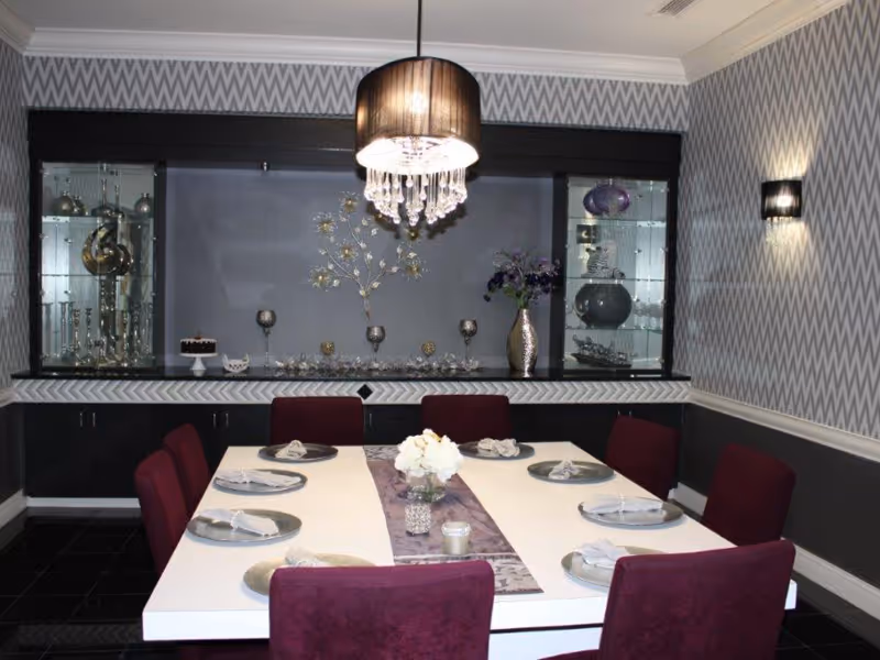 Modern dining room with a white table set for eight, burgundy chairs, a chandelier, and decorative shelving and patterned wallpaper behind.