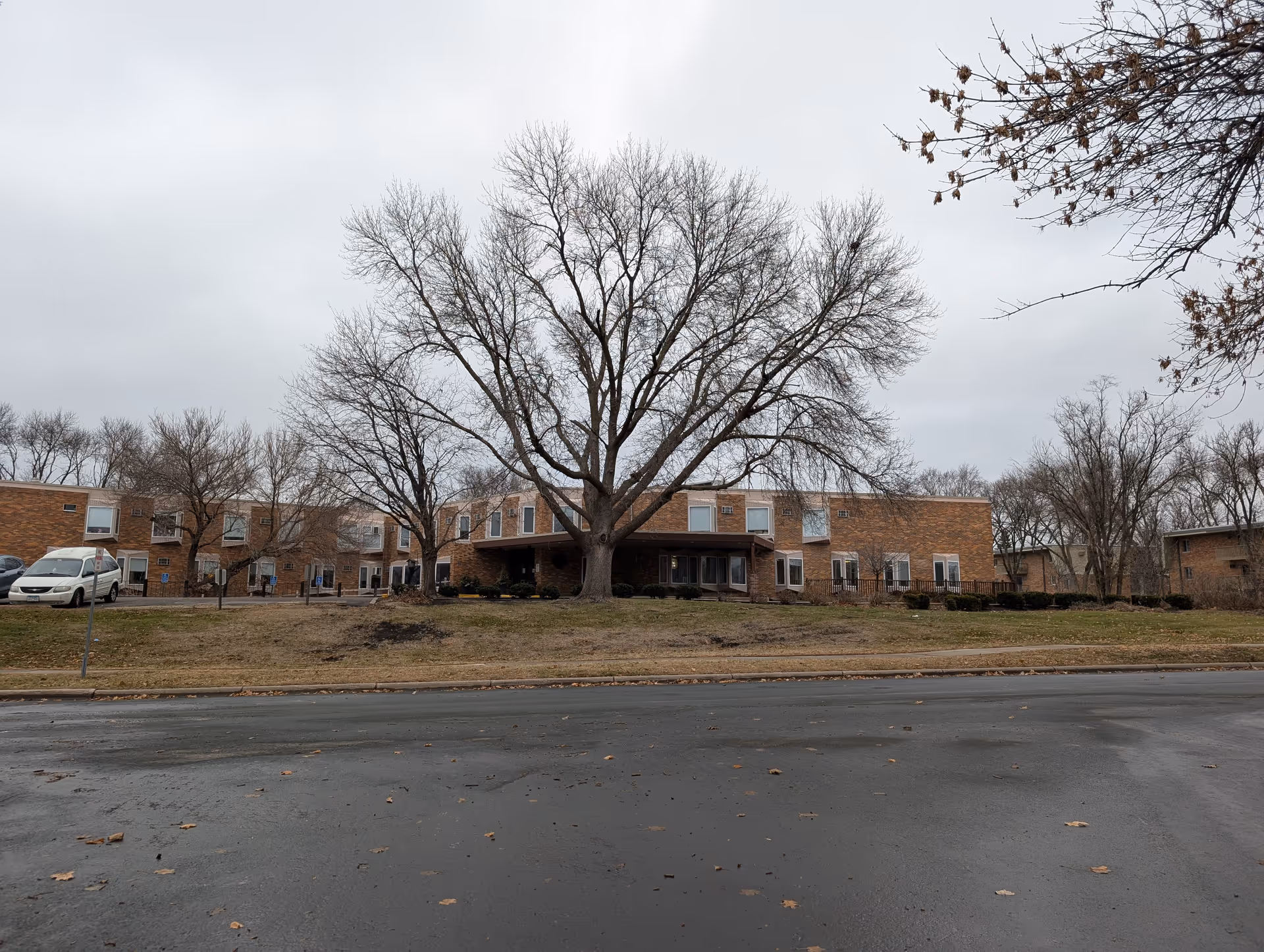 Exterior view of a two-story brick building with multiple windows, surrounded by leafless trees and a grassy area. A paved road is in the foreground under an overcast sky.