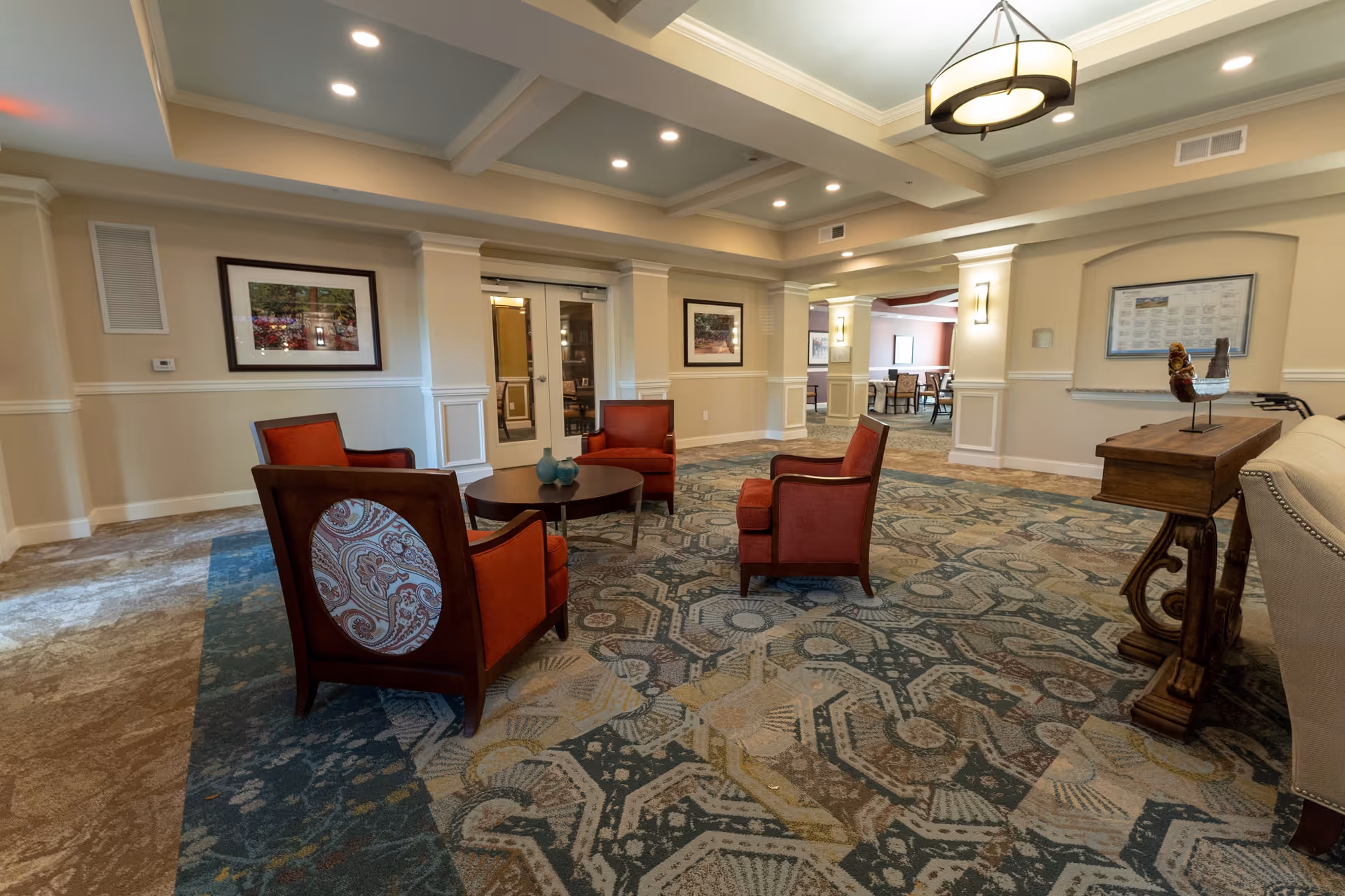 A spacious and well-lit common area with four red upholstered chairs arranged around a round wooden coffee table on a patterned carpet. The room features beige walls with white trim, framed artwork, recessed ceiling lights, and a decorative ceiling light fixture. There is a wooden console table with decorative items on the right side and a doorway leading to another room in the background.