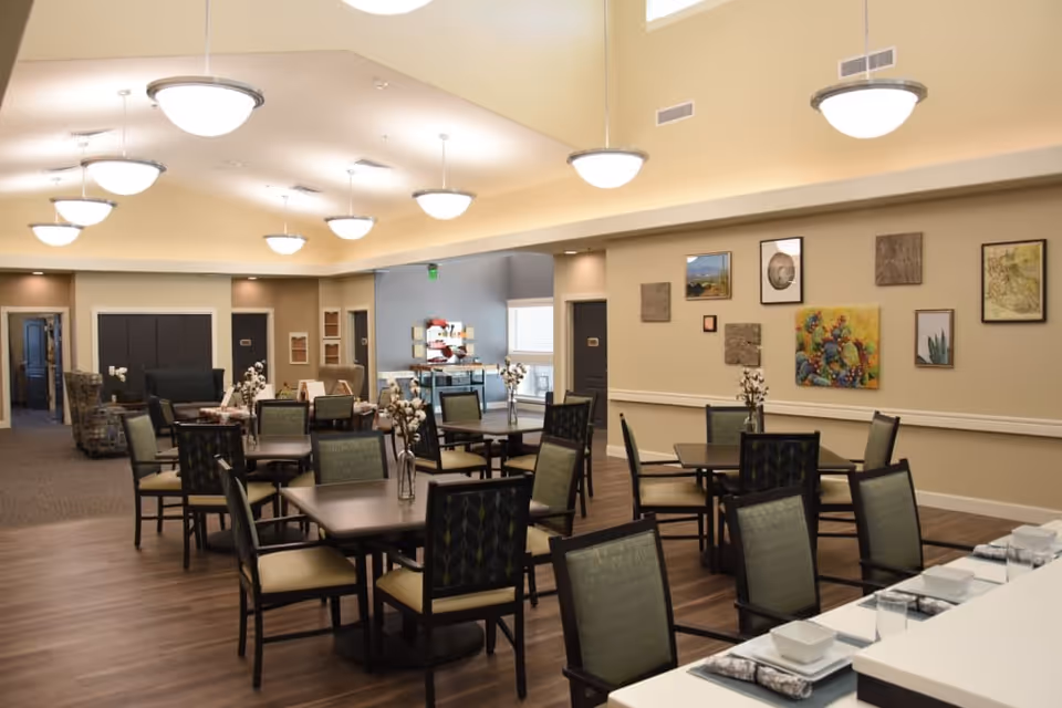 A well-lit dining room in a senior living facility with multiple square tables and chairs arranged neatly. Each table has a small vase with flowers. The room features warm beige walls adorned with various framed artworks and decorative pieces. Ceiling lights hang evenly spaced, illuminating the space. In the background, there is a seating area with armchairs and a hallway leading to other rooms.