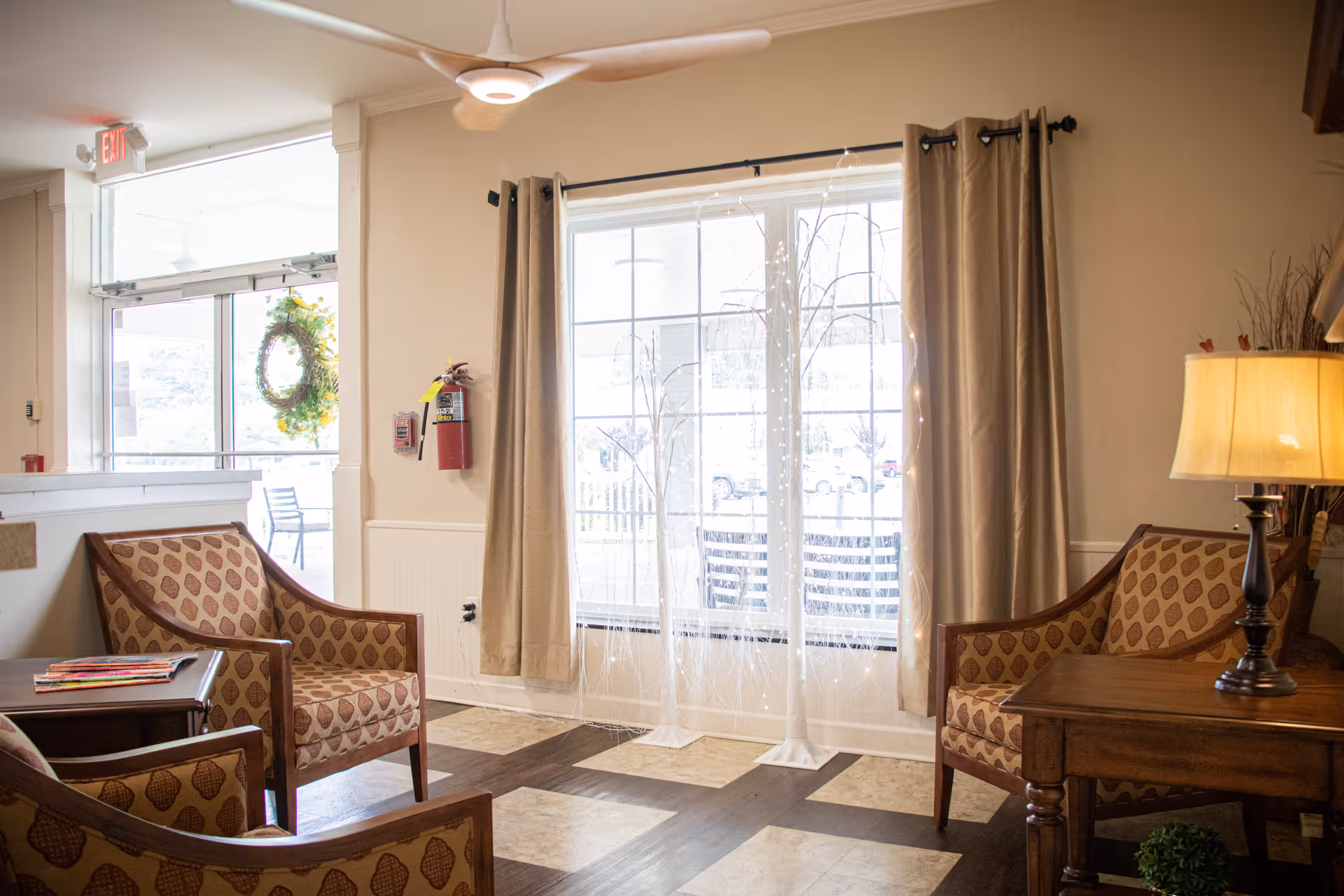 A cozy seating area in a senior living facility with three patterned armchairs arranged around a wooden side table with a lamp. Large windows with beige curtains allow natural light to fill the room. A ceiling fan is mounted above, and a fire extinguisher is visible on the wall near the entrance door decorated with a wreath.